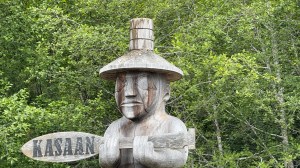 A carved figure with a Hida native hat holding a carved paddle with the word KASAAN that points to the way into town.